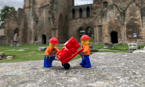 Two lego figures on a wall with a trolley of building bricks in the grounds of a cathedral 