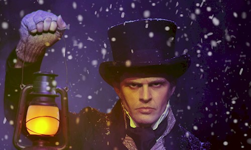 A man dressed as Ebeneezer Scrooge looking sternly at the camera holding up a lantern