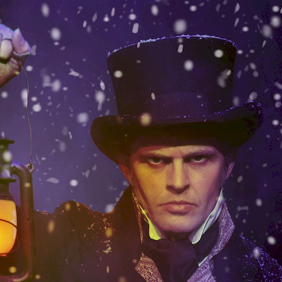 A man dressed as Ebeneezer Scrooge looking sternly at the camera holding up a lantern