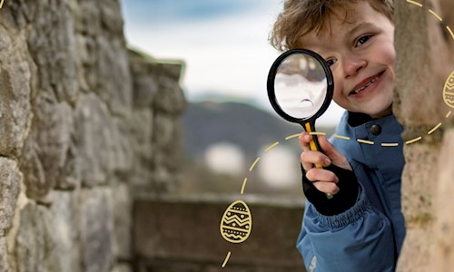 Child in a blue coat peaking round a brick wall with a magnifying glass looking for Easter egg clues