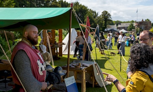A costumed performer under a canopy at a camp talking to visitors