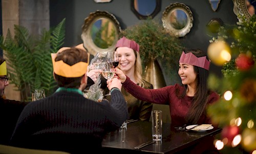 People wearing Christmas hats sitting round a table, clinking wine glasses and enjoying some food