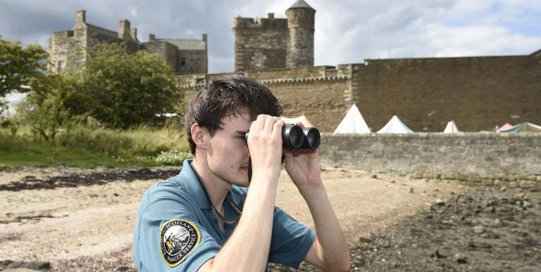 A park ranger looking out over water using binoculars with a castle in the background