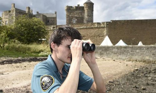 A park ranger looking out over water using binoculars with a castle in the background
