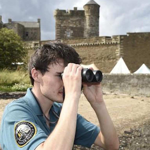 A park ranger looking out over water using binoculars with a castle in the background