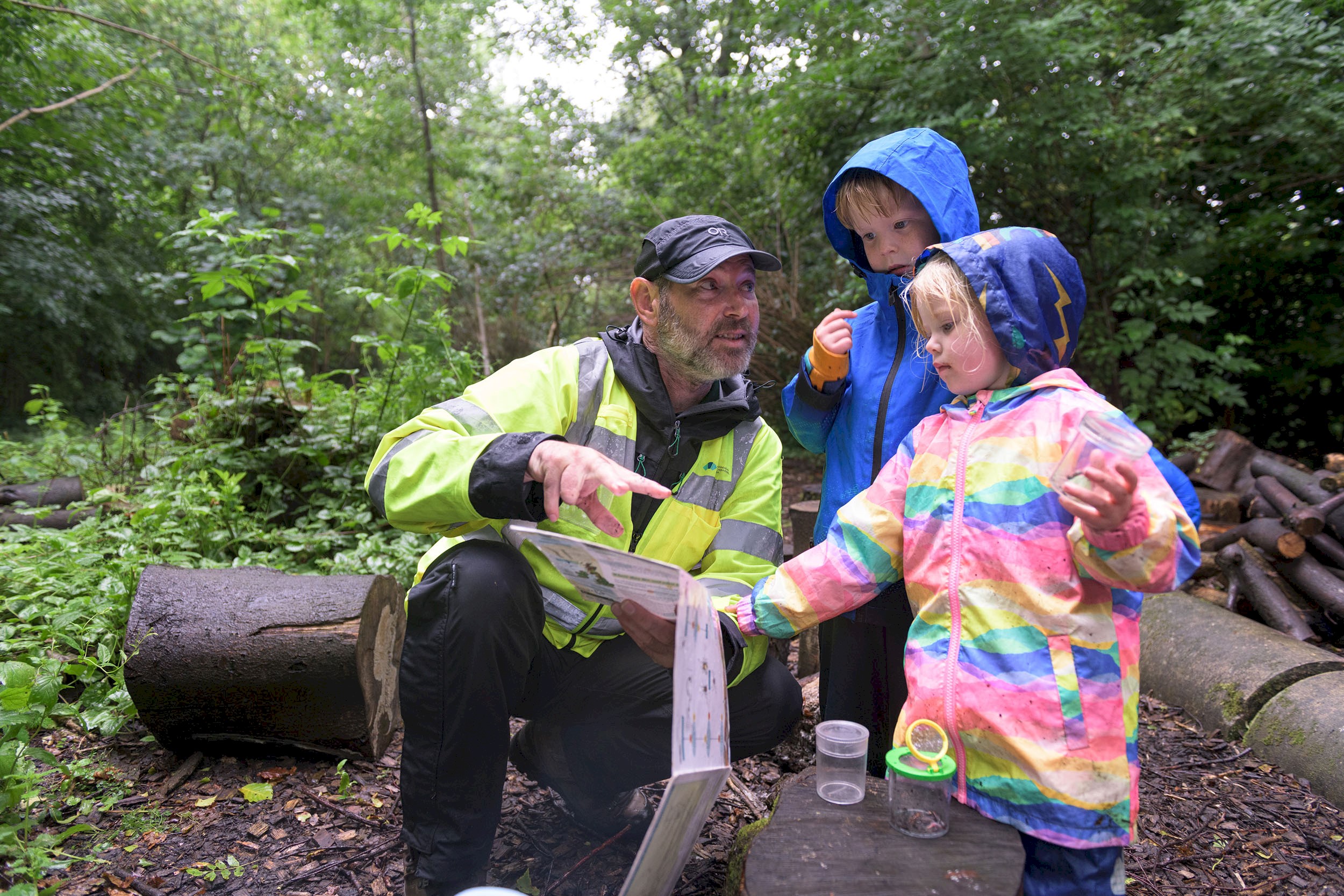 Two children with a Ranger all wearing waterproofs and looking at bugs