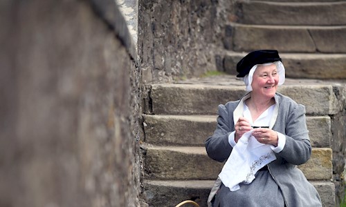A woman in period costume sitting on concrete steps doing embroidery