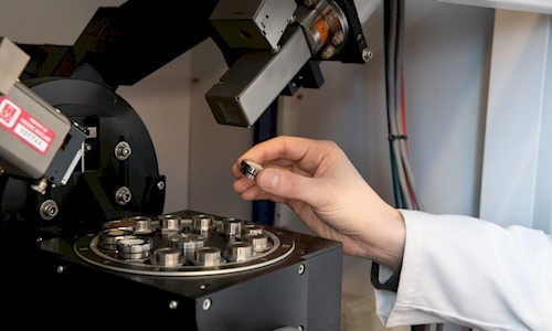 A person's hand picks up a small circular metal disc from a selection on the viewing platform of a microscope-like machine.