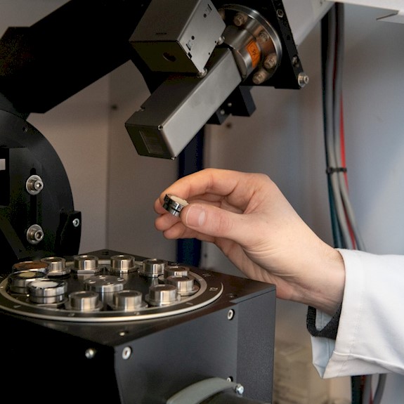 A person's hand picks up a small circular metal disc from a selection on the viewing platform of a microscope-like machine.