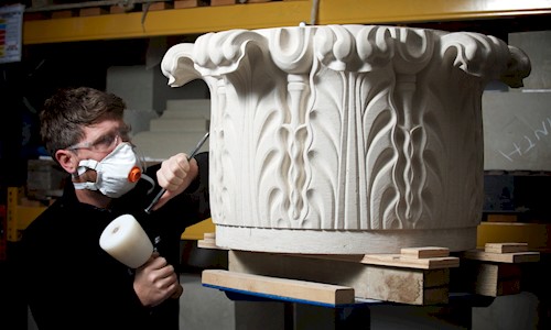 Image shows a stone mason at work, using a chisel and hammer to carve an ornate marble column piece. They are wearing protective goggles and a mask.