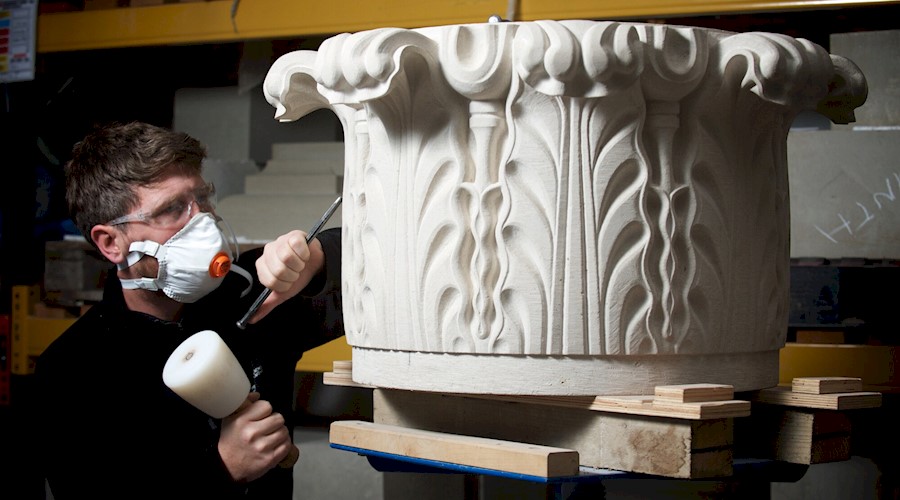 Image shows a stone mason at work, using a chisel and hammer to carve an ornate marble column piece. They are wearing protective goggles and a mask.