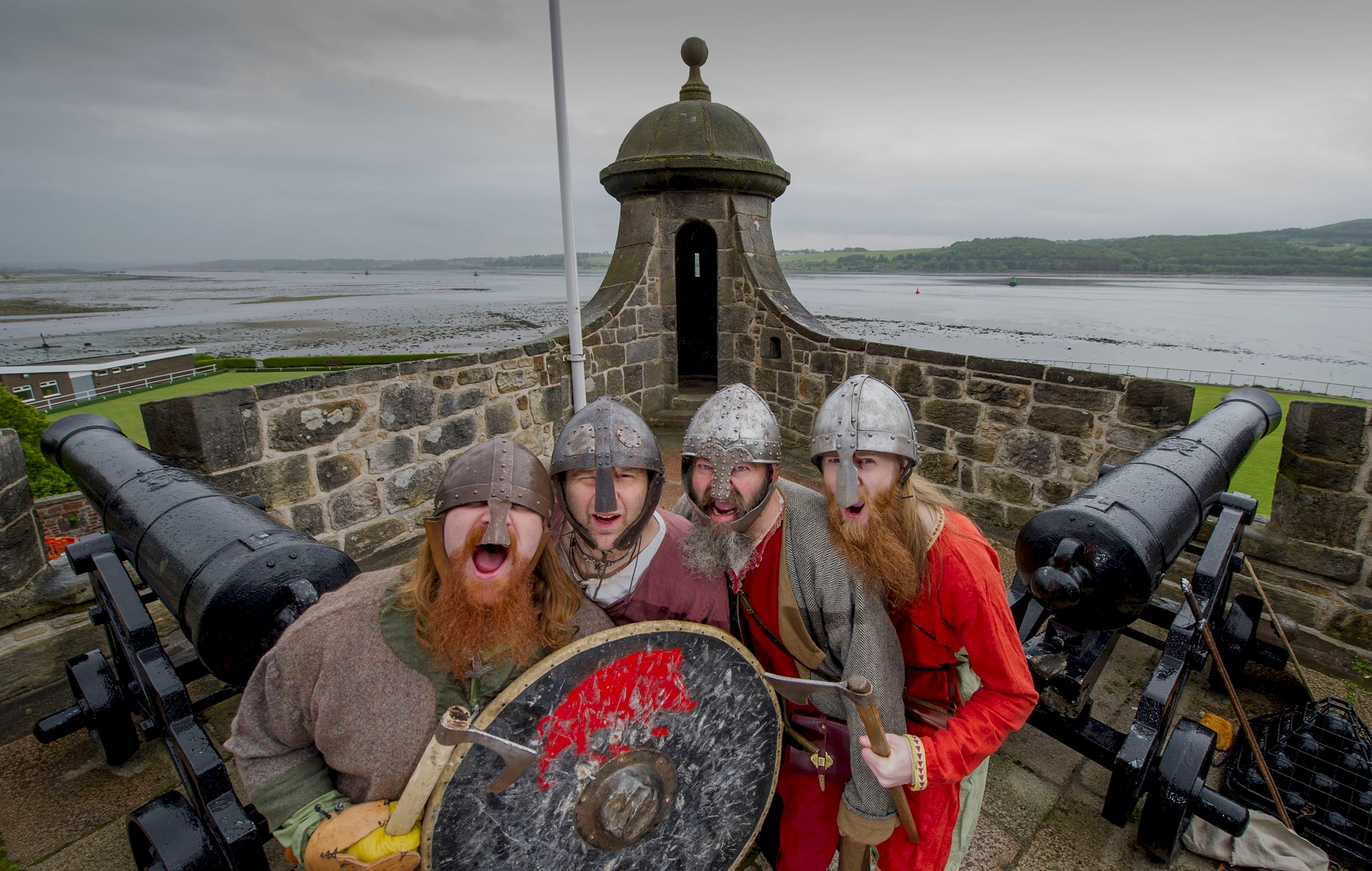 Costumed performers dressed as vikings and roaring at the camera with cannons and the Firth of Clyde in the background