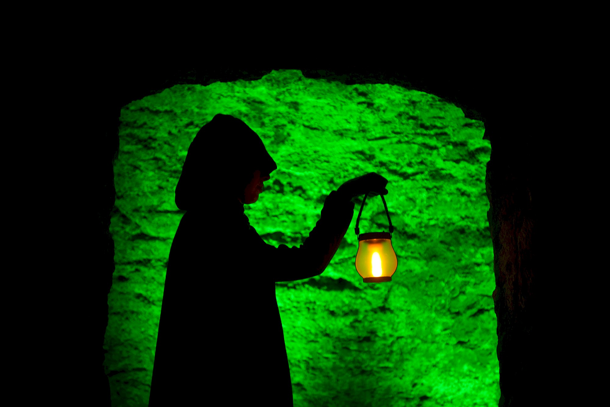 Silhouette of a hooded figure standing in a stone doorway, holding a glowing lantern. The lantern casts a warm yellow light, contrasting with the vivid green illumination on the rough stone wall behind, creating a dramatic and mysterious atmosphere.