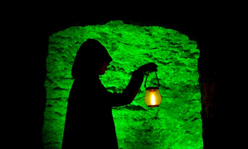 Silhouette of a hooded figure standing in a stone doorway, holding a glowing lantern. The lantern casts a warm yellow light, contrasting with the vivid green illumination on the rough stone wall behind, creating a dramatic and mysterious atmosphere.