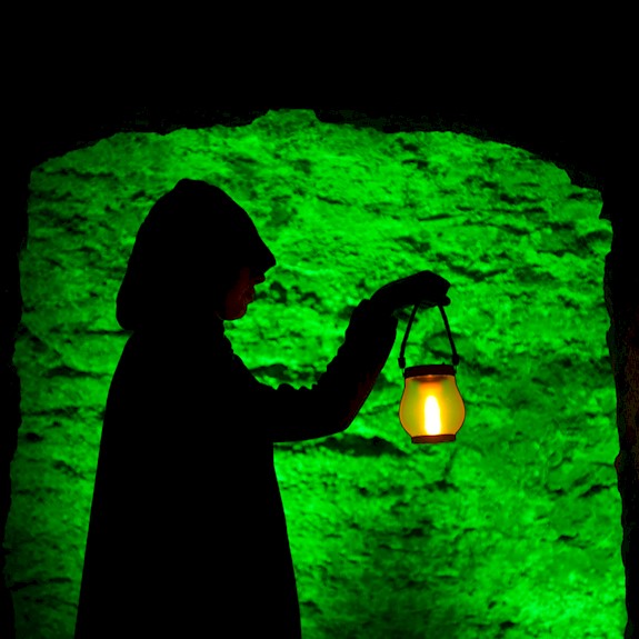 Silhouette of a hooded figure standing in a stone doorway, holding a glowing lantern. The lantern casts a warm yellow light, contrasting with the vivid green illumination on the rough stone wall behind, creating a dramatic and mysterious atmosphere.