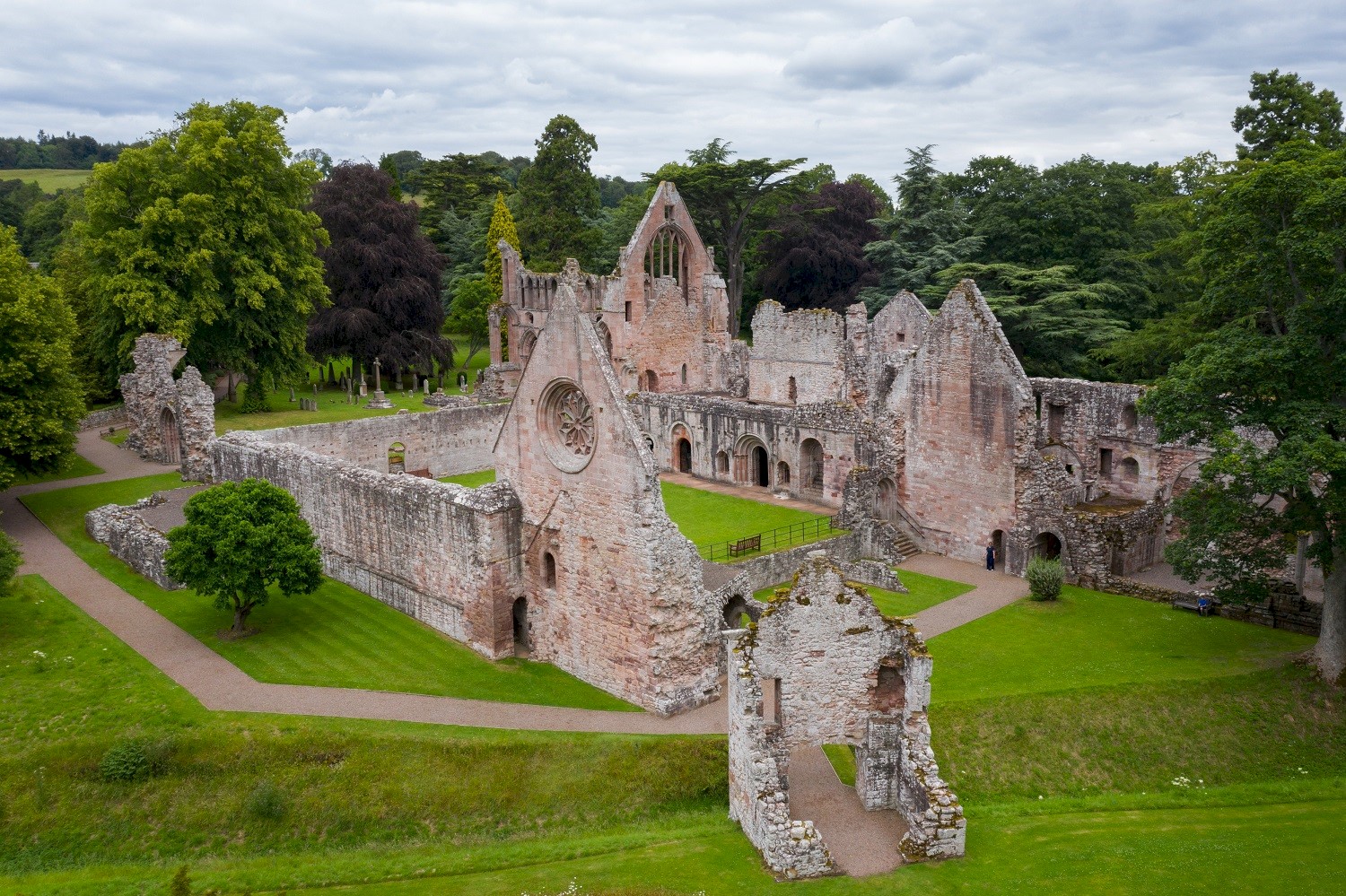 the ruins of a medieval abbey. It's a roofless structure but with significant walls still standing. It's surrounded by a grassy lawn and trees and seems peaceful.