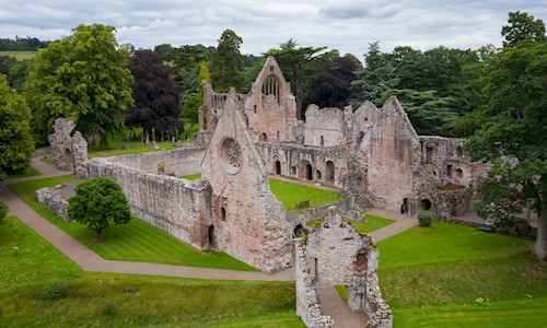 the ruins of a medieval abbey. It's a roofless structure but with significant walls still standing. It's surrounded by a grassy lawn and trees and seems peaceful.