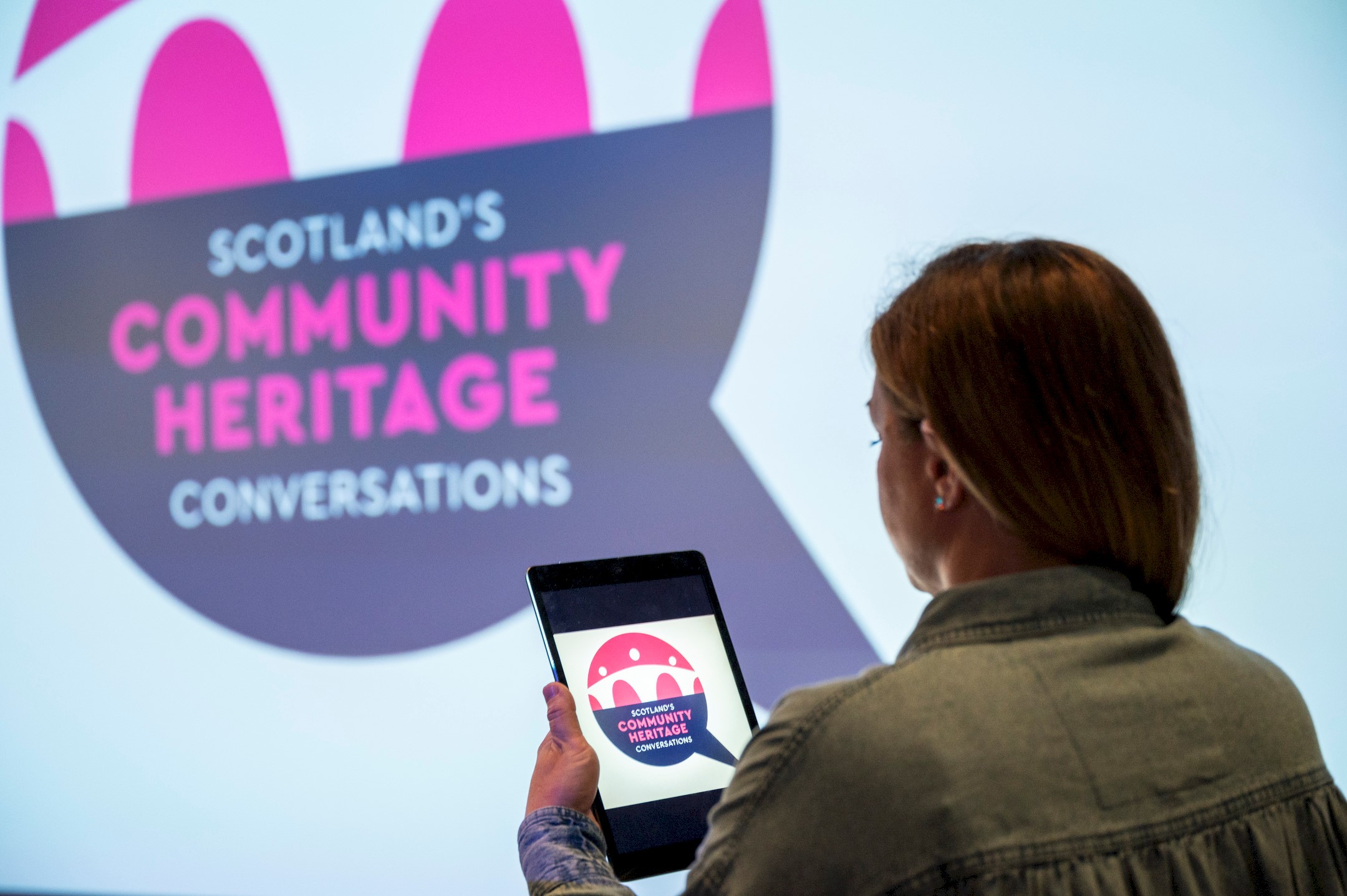 A person holding a tablet standing in front of a big screen at an exhibition centre. A blue, white and pink logo reading "Scotland's Community Heritage Conversations" is on both the small tablet screen and the big screen.
