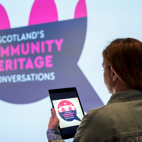 A person holding a tablet standing in front of a big screen at an exhibition centre. A blue, white and pink logo reading "Scotland's Community Heritage Conversations" is on both the small tablet screen and the big screen.