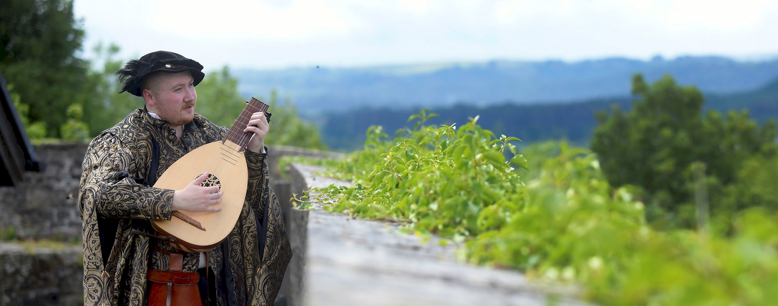 A man in period costume playing a lute while looking over a wall with lovely views of greenery and hills