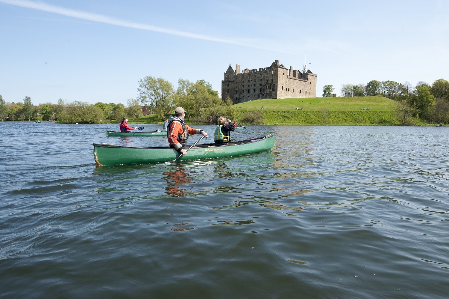 Three people in a canoe paddle across Linlithgow Loch with Linlithgow Palace in the background