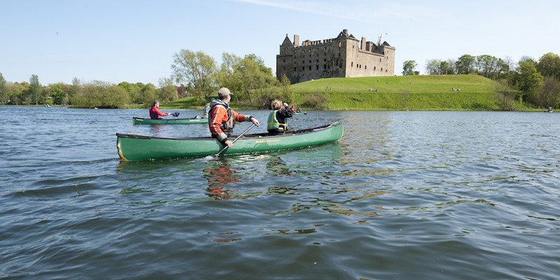 Three people in a canoe paddle across Linlithgow Loch with Linlithgow Palace in the background