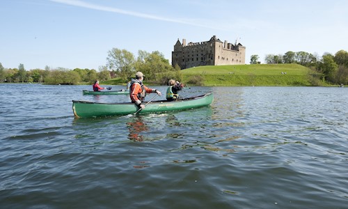 Three people in a canoe paddle across Linlithgow Loch with Linlithgow Palace in the background