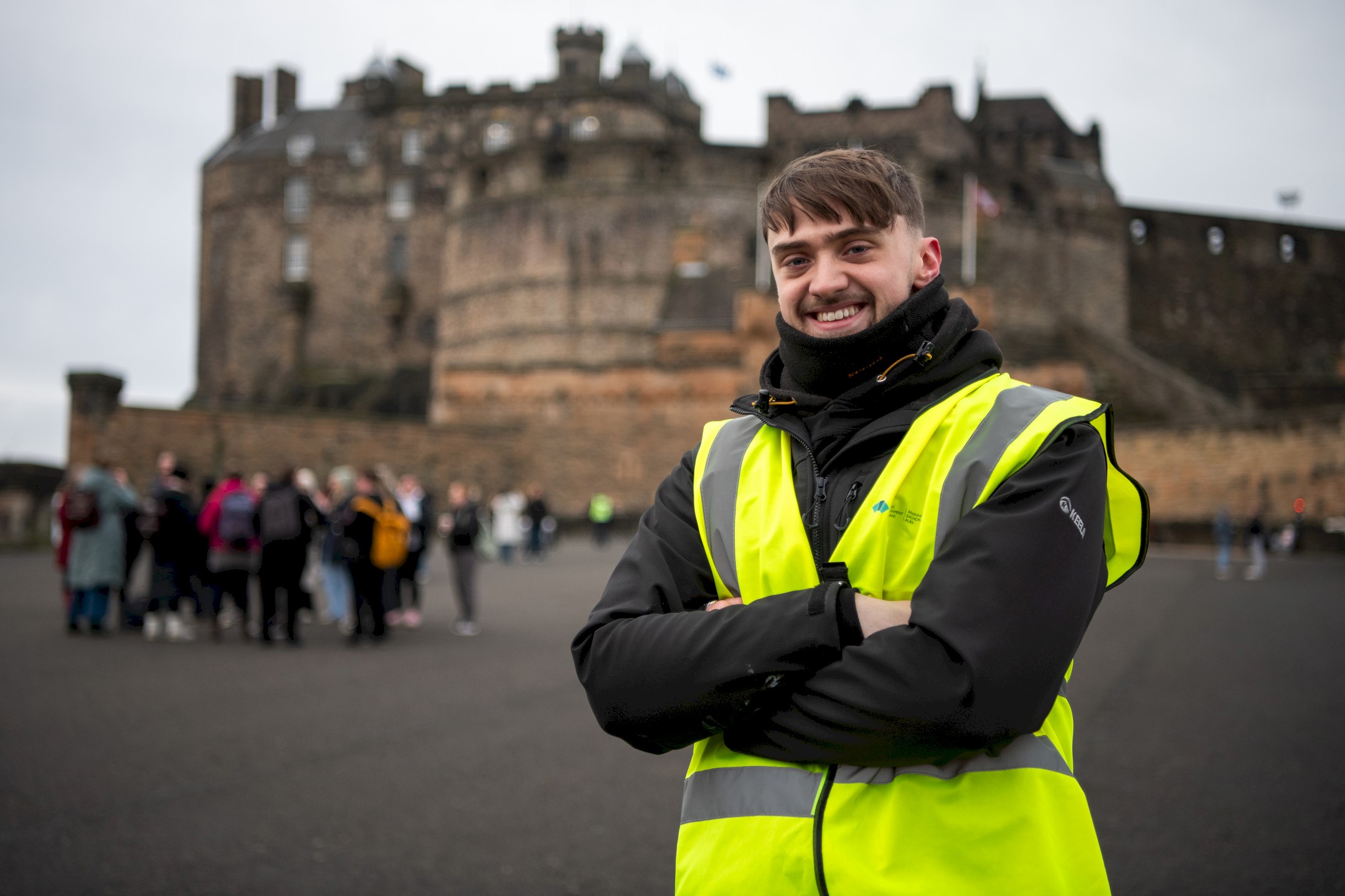 Person wearing a high-visibility yellow vest and dark jacket stands with arms crossed in the foreground of Edinburgh Castle.