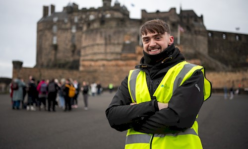 Person wearing a high-visibility yellow vest and dark jacket stands with arms crossed in the foreground of Edinburgh Castle.