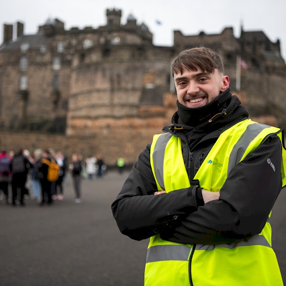 Person wearing a high-visibility yellow vest and dark jacket stands with arms crossed in the foreground of Edinburgh Castle.