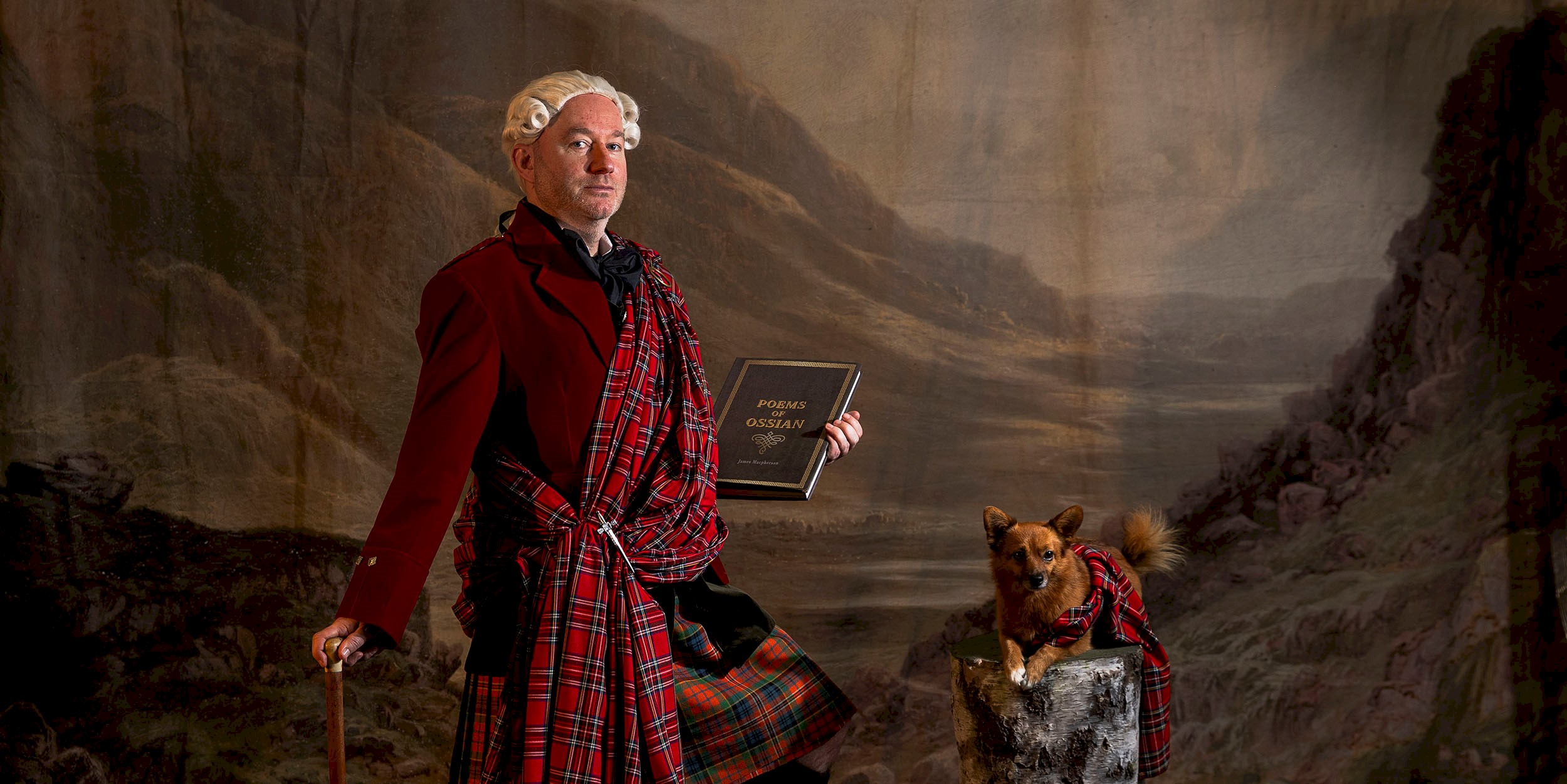 A man in a kilt with a sash holding a book and a walking stick, alongside is a small dog lying on a log with a tartan sash over it