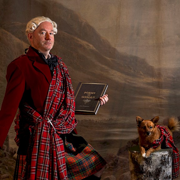 A man in a kilt with a sash holding a book and a walking stick, alongside is a small dog lying on a log with a tartan sash over it