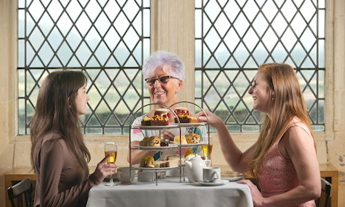 three people sitting at a table with a variety of sandwiches and cakes on a cake stand along with glasses of prosecco and tea in cups and saucers