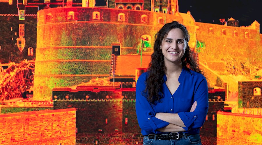 A person stands smiling with their arms folded in front of a large wall-displayed computer generated image of Edinburgh Castle.