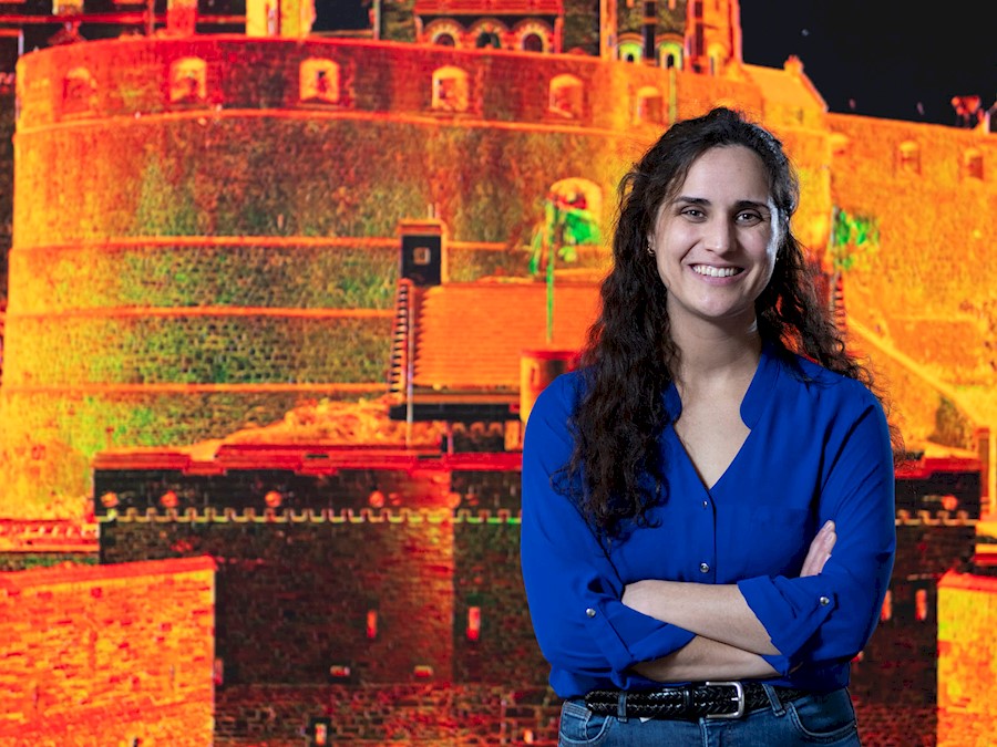 A person stands smiling with their arms folded in front of a large wall-displayed computer generated image of Edinburgh Castle.