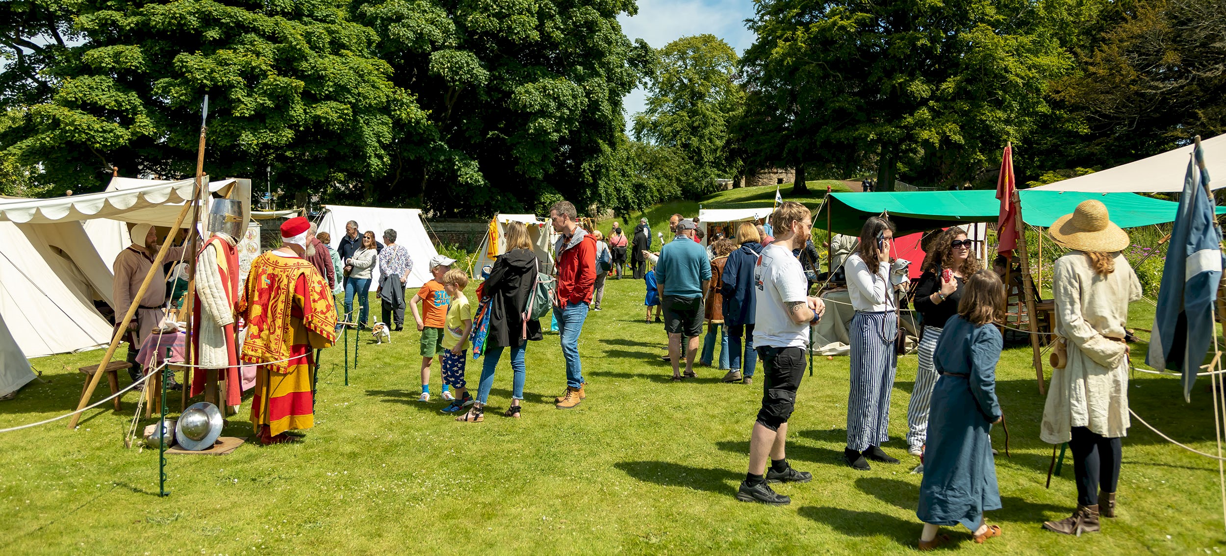 Visitors walking round a medieval encampment in the glorious sunshine talking to the tradespeople at their stalls 