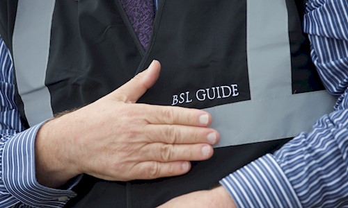 A BSL tour guide holding their hand flat to their chest where the jacket is embroidered with BSL Guide