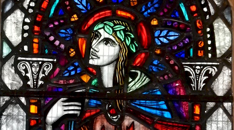 A multicoloured stained glass window featuring a design of a person with long hair looking to the left. They are wearing a cloak fastened with a red jewel and have a band of leaves in their hair.