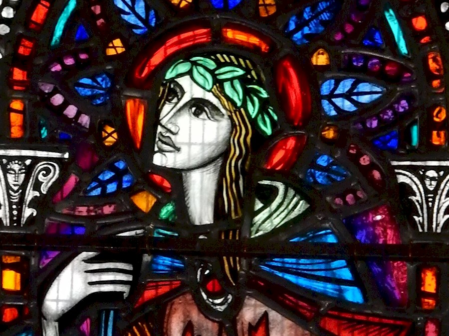 A multicoloured stained glass window featuring a design of a person with long hair looking to the left. They are wearing a cloak fastened with a red jewel and have a band of leaves in their hair.