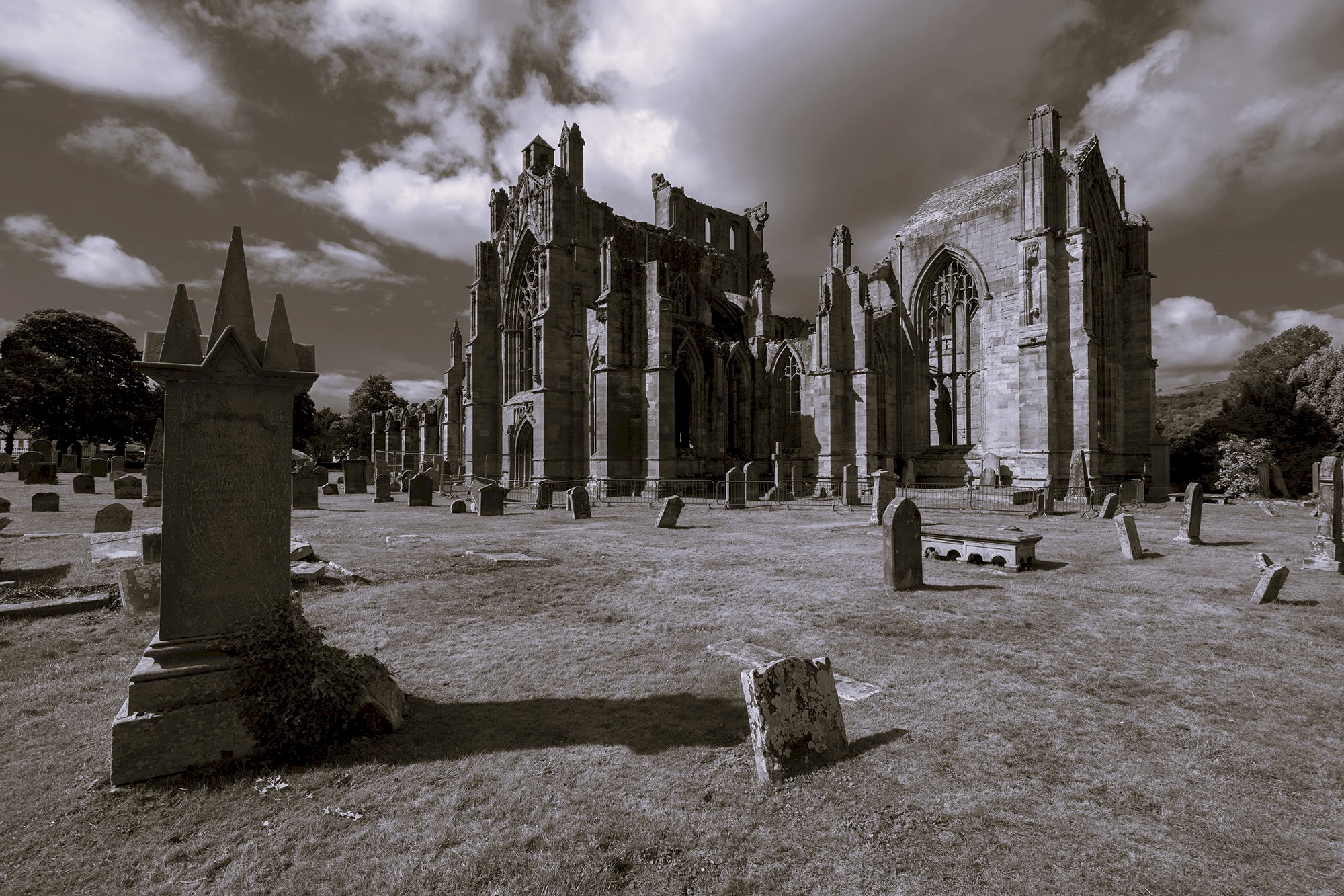 Black-and-white photograph of a large Gothic abbey ruin surrounded by an historic graveyard. Weathered headstones stand across the grass in the foreground, while the roofless stone structure with tall arched windows dominates the centre of the image. Dramatic clouds fill the sky above, adding a striking and atmospheric quality to the scene.