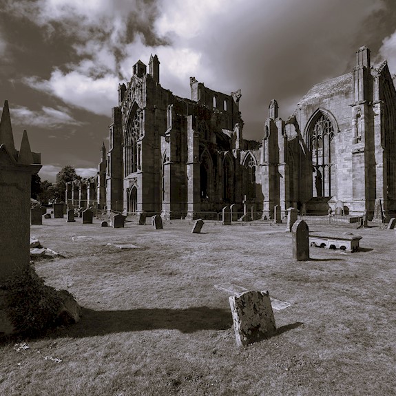 Black-and-white photograph of a large Gothic abbey ruin surrounded by an historic graveyard. Weathered headstones stand across the grass in the foreground, while the roofless stone structure with tall arched windows dominates the centre of the image. Dramatic clouds fill the sky above, adding a striking and atmospheric quality to the scene.