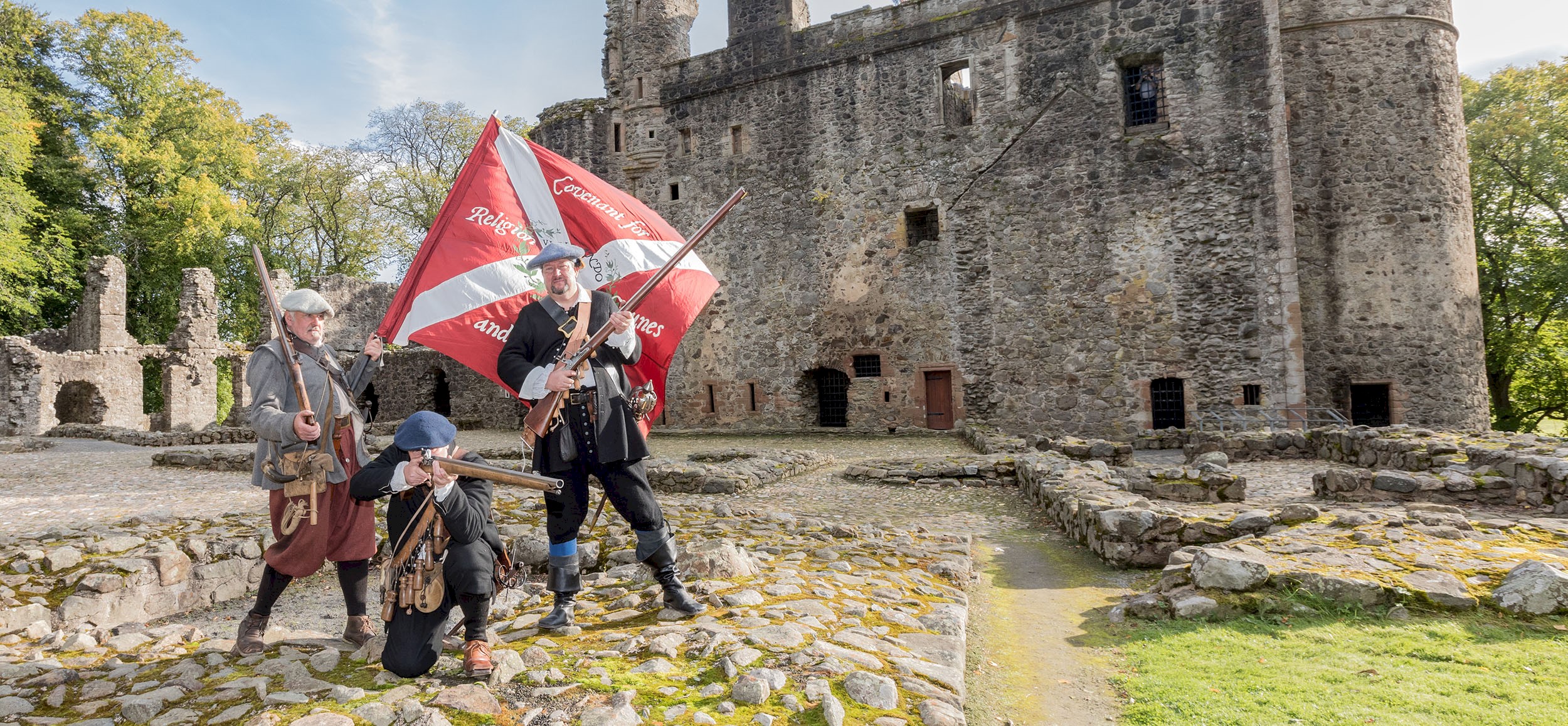 Three costumed performers holding muskets and standing in the grounds of a castle