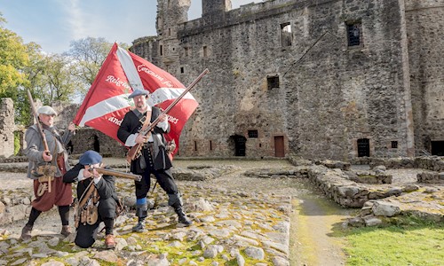 Three costumed performers holding muskets and standing in the grounds of a castle