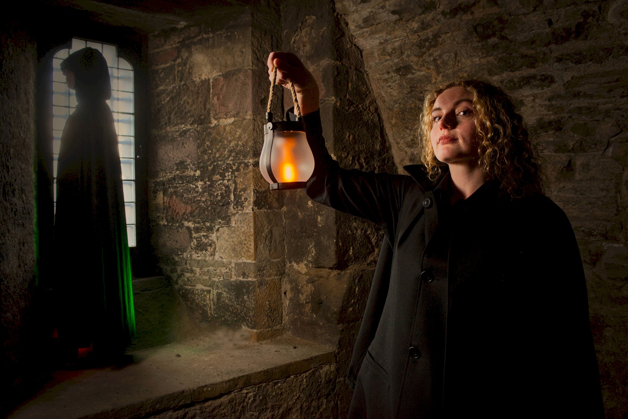 A person in a dark coat holds a glowing lantern inside a stone-walled chamber. In the background, a hooded figure stands silhouetted beside a tall leaded-glass window, while the lantern casts warm light across the textured stone walls, creating a dramatic and atmospheric scene.