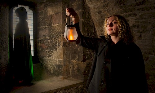 A person in a dark coat holds a glowing lantern inside a stone-walled chamber. In the background, a hooded figure stands silhouetted beside a tall leaded-glass window, while the lantern casts warm light across the textured stone walls, creating a dramatic and atmospheric scene.