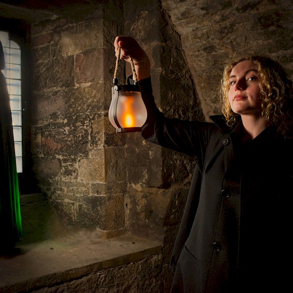 A person in a dark coat holds a glowing lantern inside a stone-walled chamber. In the background, a hooded figure stands silhouetted beside a tall leaded-glass window, while the lantern casts warm light across the textured stone walls, creating a dramatic and atmospheric scene.