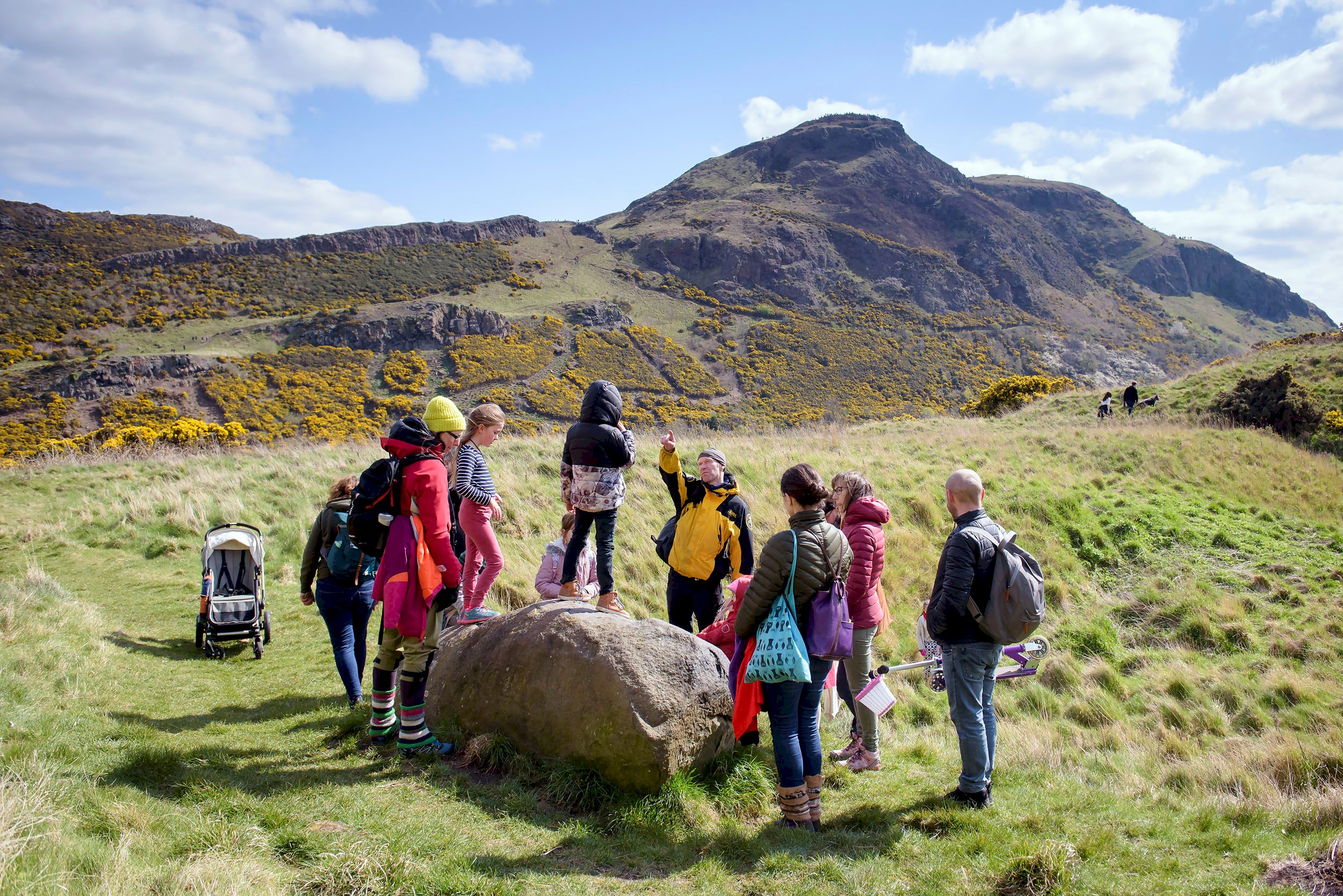 A group of people surrounding a park Ranger listening to them giving information about Holyrood park