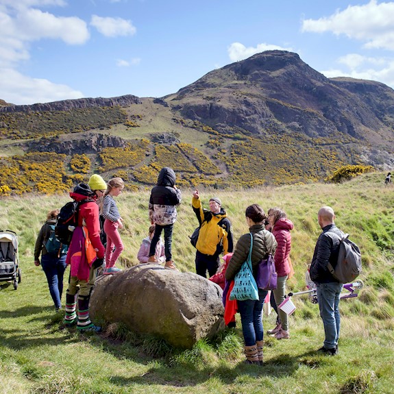 A group of people surrounding a park Ranger listening to them giving information about Holyrood park