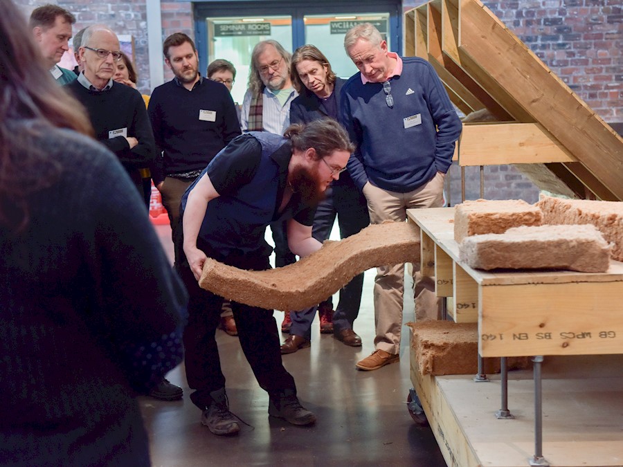 Image shows a person pulling a section of loft insulation from a display case with others watching in the background.