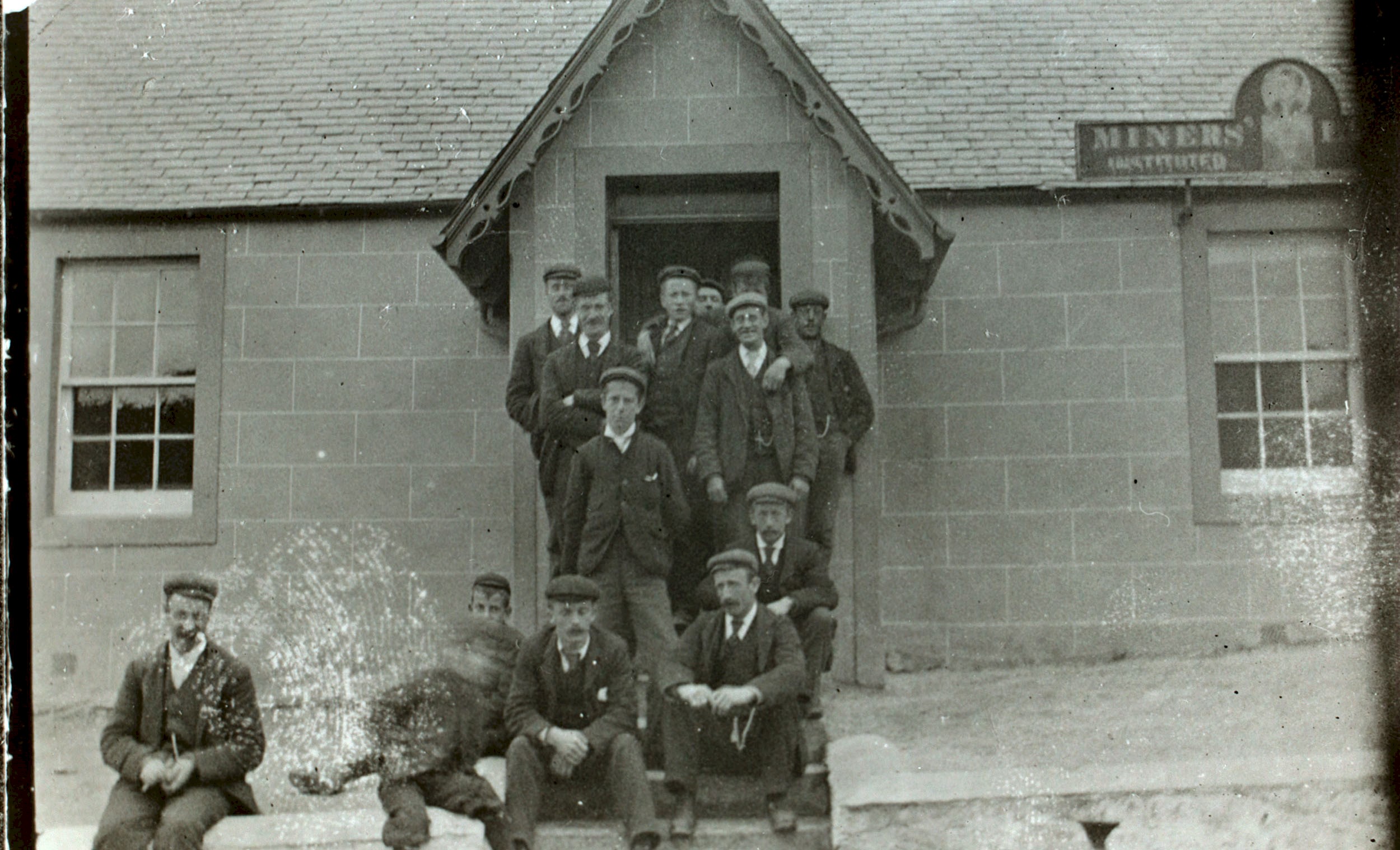 A black and white photo shows 14 men dressed in suits with black boots and flat caps. They sit and stand on the steps of a stone building with a slate roof.